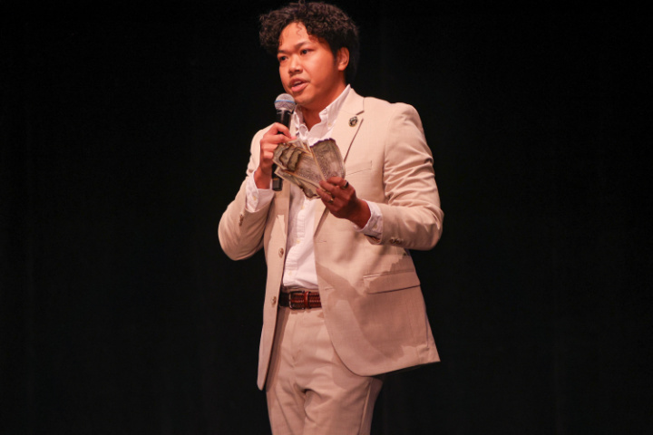 Leland Ramirez, founder and CEO of VroomBrick, presents on stage during the Virginia Cup startup pitch competition, holding cash while explaining how the company helps make homeownership more affordable.
