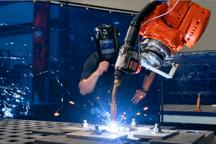 A person wearing a welding helmet watches an orange robotic arm deposit metal on a worktable, producing bright sparks.