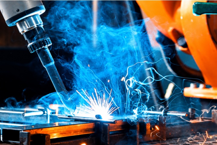 Robotic welding equipment produces bright sparks and blue smoke while working on a metal component.
